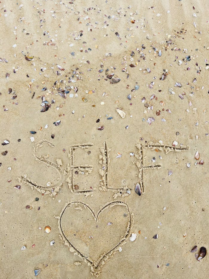The word 'self' with a heart shape drawn in sand amidst scattered seashells on a beach.